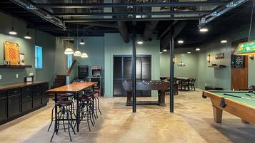 A spacious room with a pool table, foosball table, and bar area. There are multiple seating options and a staircase leading to an upper level.