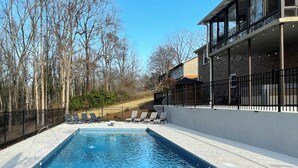 A rectangular swimming pool with a tiled edge, surrounded by a black metal fence. There are lounge chairs along the poolside, and a building with large windows and a balcony is adjacent to the pool. - Luxury 6,000+ sq ft Hideaway with Heated Pool, Spa, & Sauna - near Greenville (Taylors)