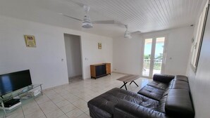 A living room with a leather sofa, a TV on a glass stand, a ceiling fan, and a sliding glass door leading to a balcony.
