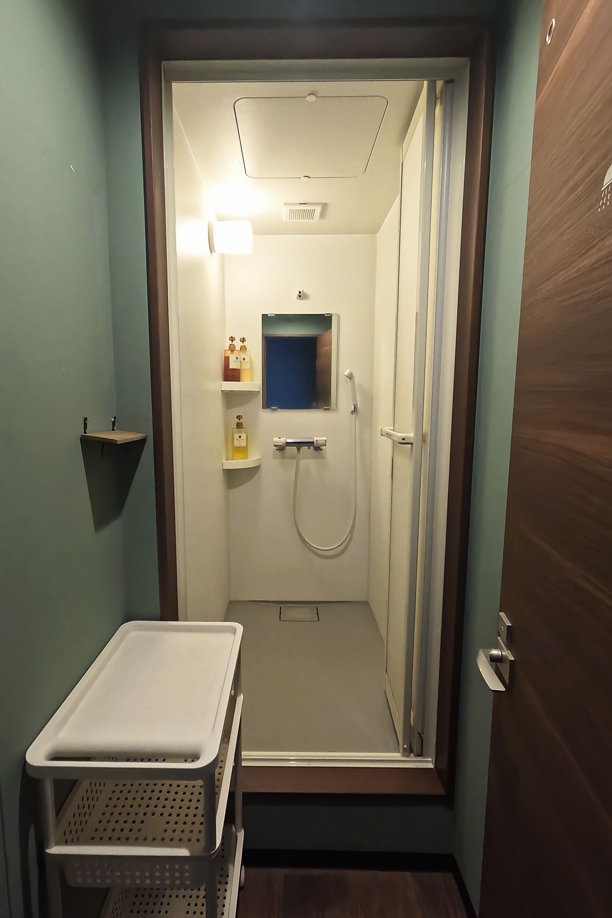 A small, enclosed bathroom with a shower, a sink, and a mirror. There are shelves with toiletries and a small shelf on the wall.