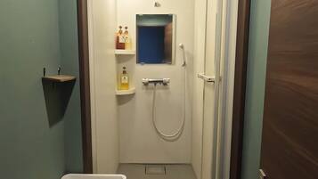 A small, enclosed bathroom with a shower, a sink, and a mirror. There are shelves with toiletries and a small shelf on the wall.