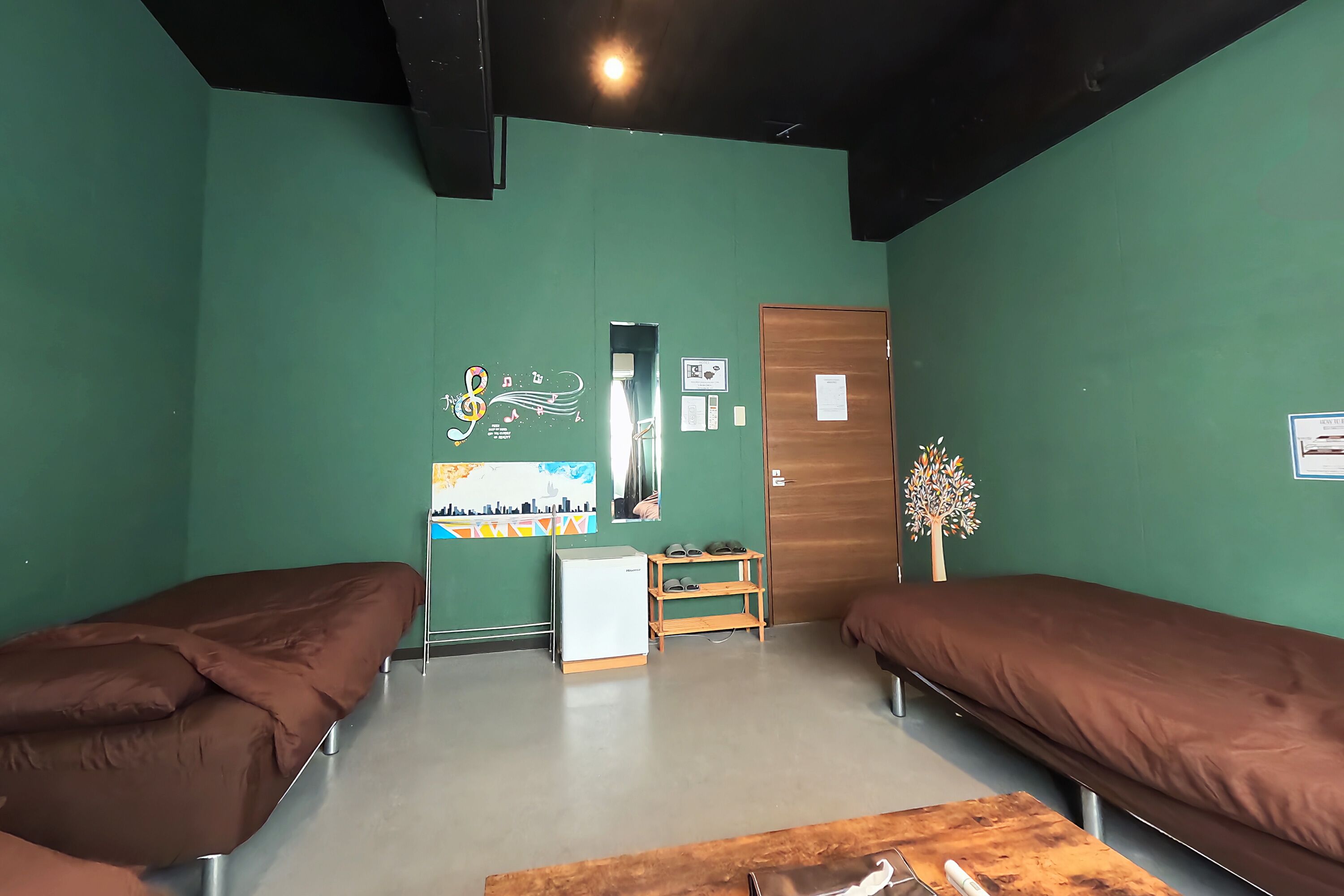 Une chambre avec deux lits simples, une petite table, un réfrigérateur et une étagère en bois où sont rangées des chaussures.