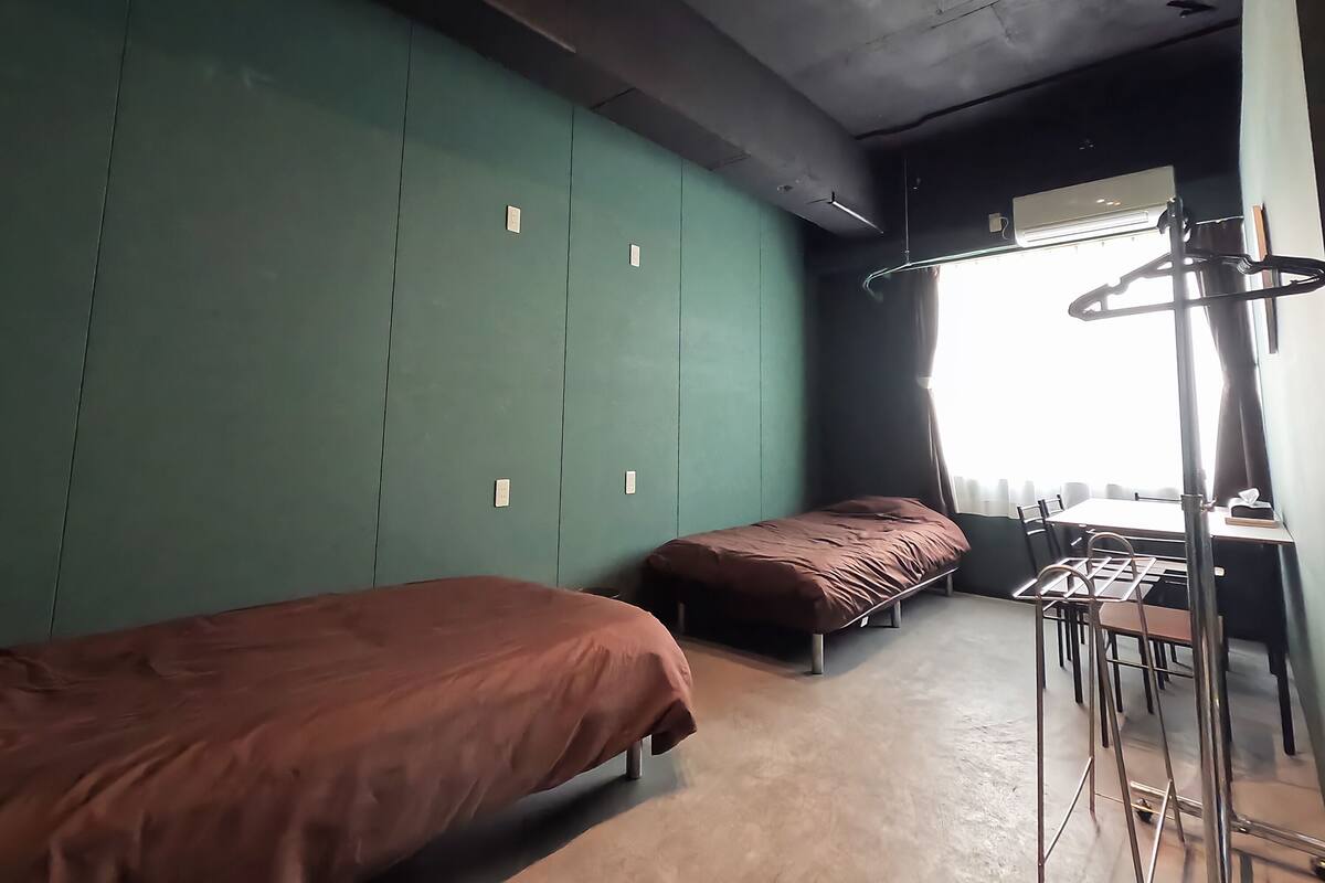 A room with two single beds, a desk with chairs, and a large window with curtains.