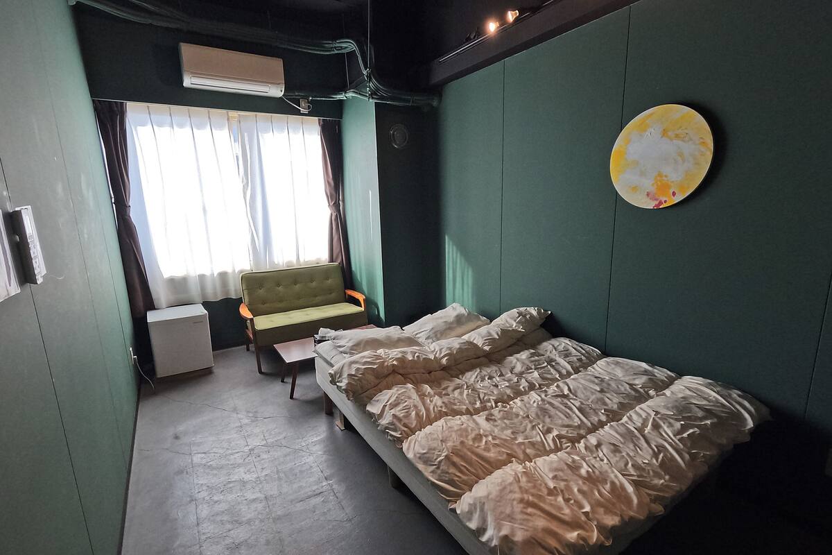 A small, neatly arranged room with a bed, a chair, a small table, and a window with curtains.