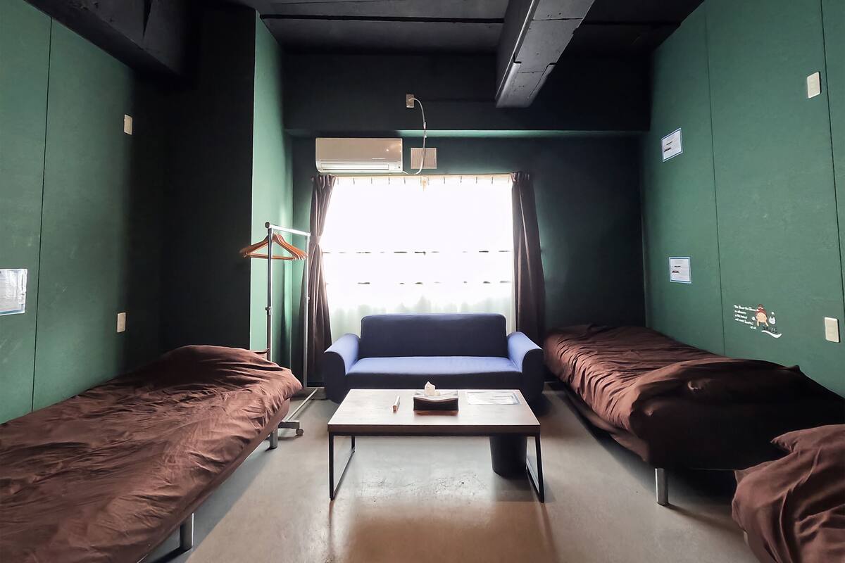 A room with two single beds, a small table, a window with curtains, and an air conditioning unit.