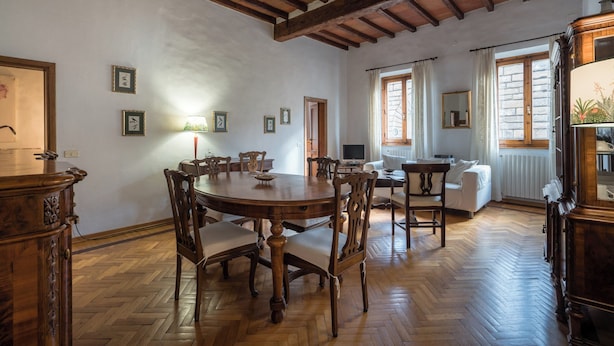 A spacious room with a wooden dining table, chairs, and a sofa. There are framed pictures on the walls and a lamp on the table.
