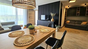 A modern living room with a wooden dining table, black chairs, a grey sofa, and a TV mounted on the wall. - Apartament Nefryt - 365PAM (Sarbinowo)