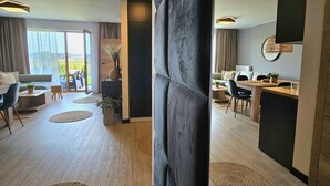 A modern living room with a sofa, coffee table, and a dining area with a table and chairs. There is a large window providing natural light and a view of the outdoors. - Apartament Nefryt - 365PAM (Sarbinowo)