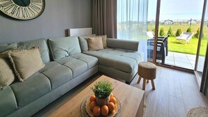 A modern living room with a sectional sofa, a round table with a plant and oranges, and a large window with a view of a green landscape. - Apartament Nefryt - 365PAM (Sarbinowo)