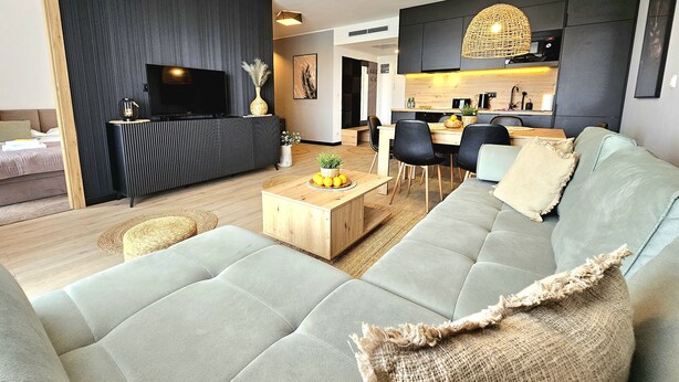 A modern living room with a sectional sofa, a coffee table, and a TV. There is a kitchen area with a dining table and chairs. - Apartament Nefryt - 365PAM (Sarbinowo)