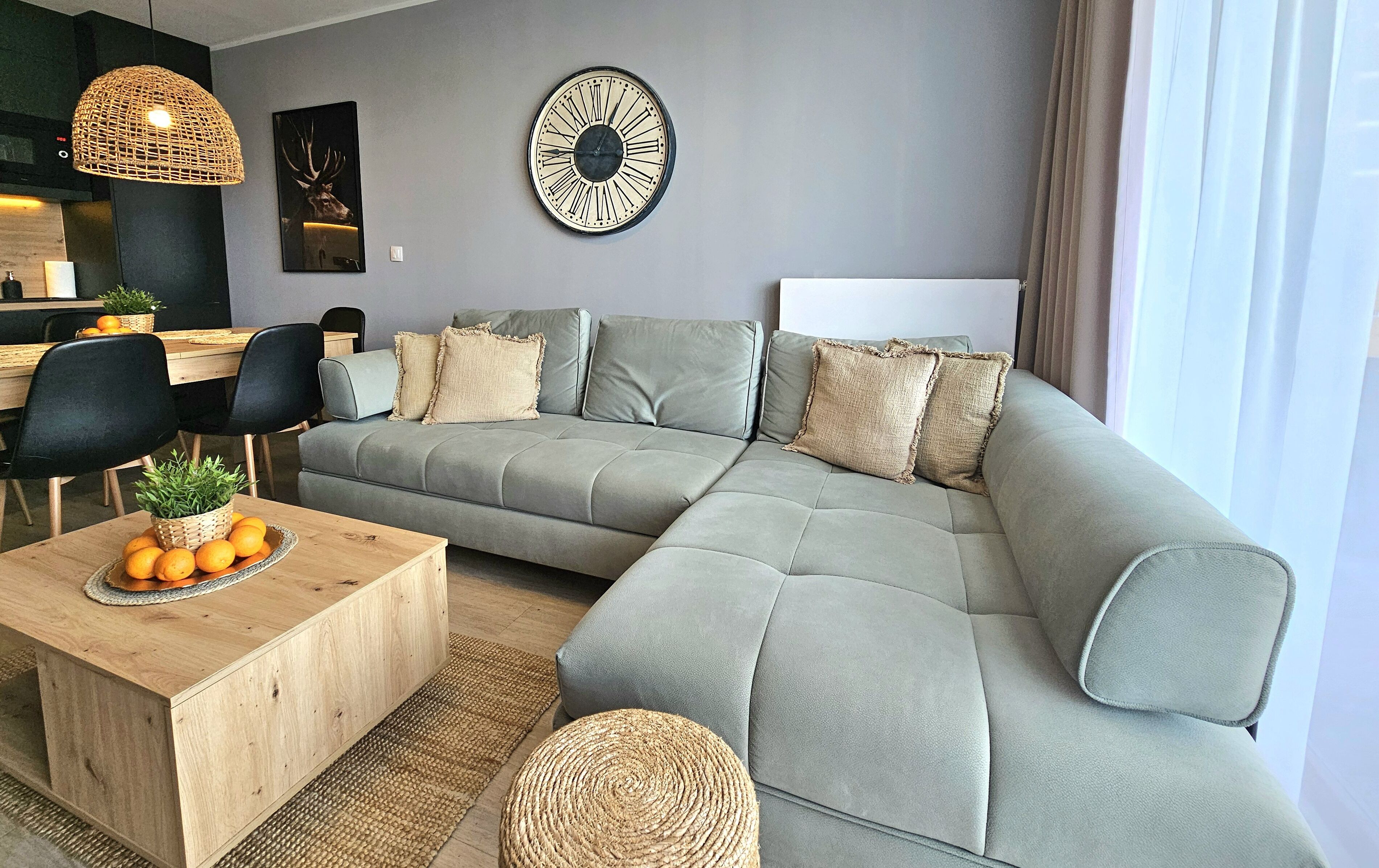 Ein modernes Wohnzimmer mit einem grauen Ecksofa, einem Holztisch mit Pflanze und Orangen, schwarzen Stühlen, einer gewobenen Pendelleuchte und einer Wanduhr.