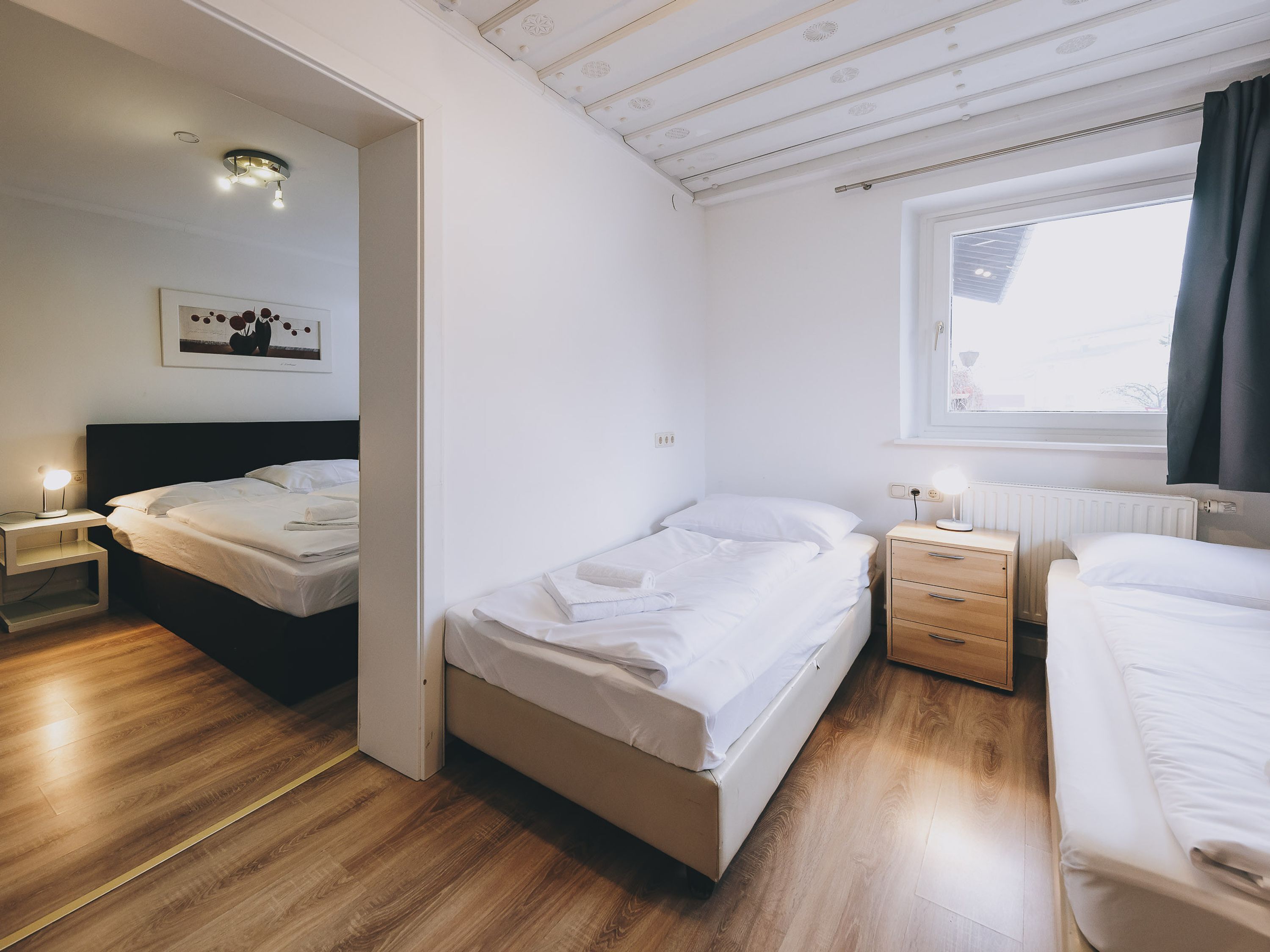 A bedroom with two beds, a bedside table with a lamp, a radiator, and a window with curtains.