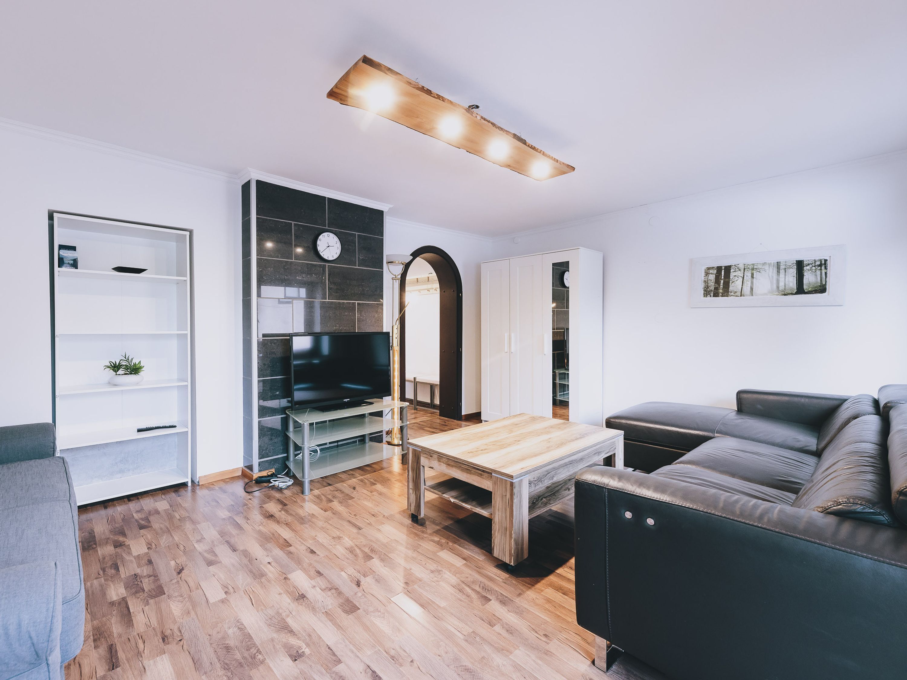 A modern living room with a wooden coffee table, a black leather sofa, a TV on a stand, a bookshelf, and a wardrobe.