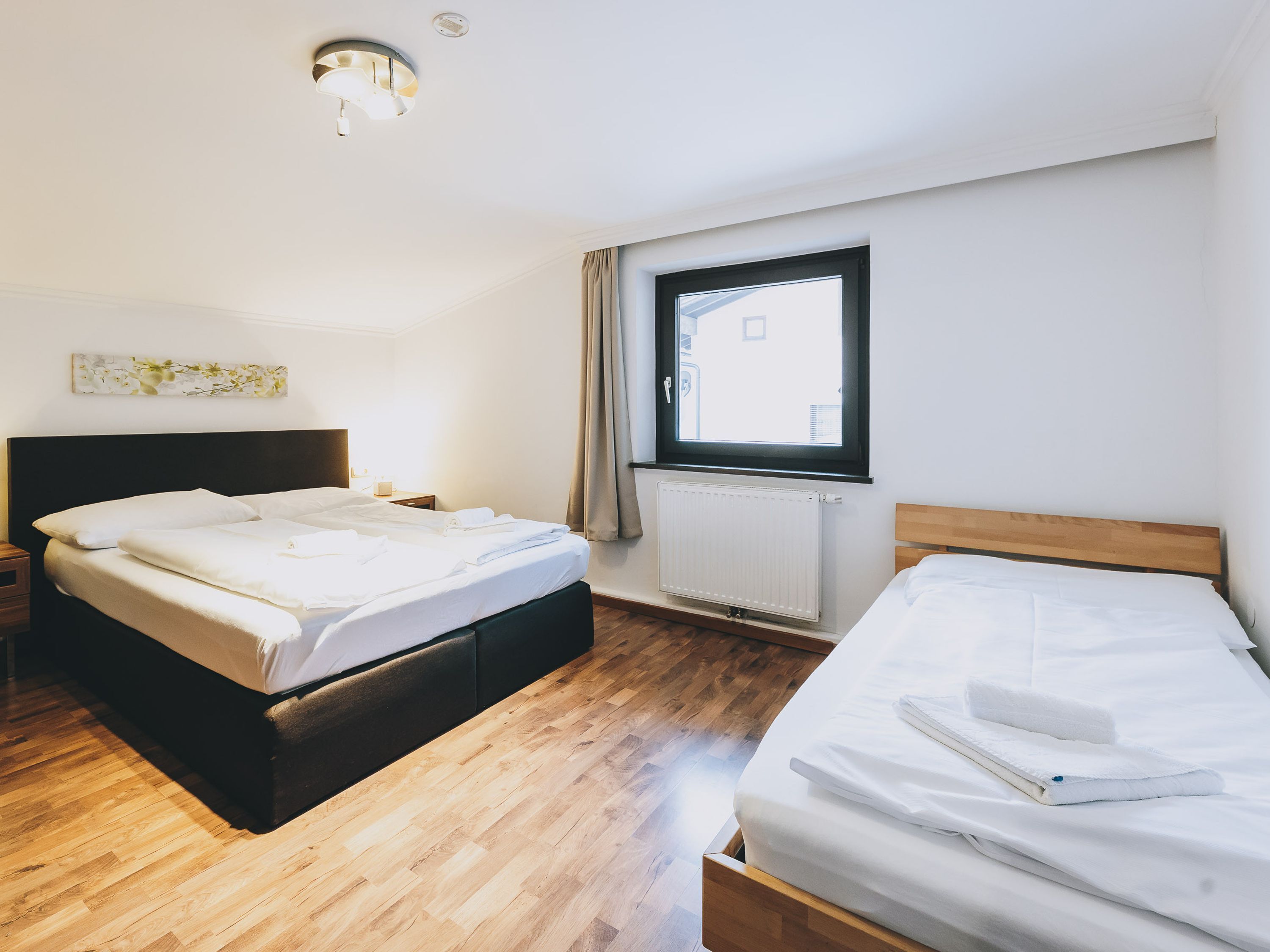 A hotel room with two beds, a wooden floor, a window with curtains, a radiator, and a ceiling light fixture.