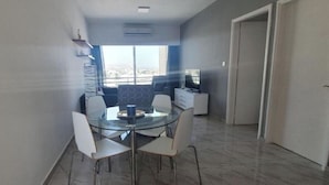 A modern dining area with a glass table and four chairs, a flat-screen TV, and a view of the city through large windows. - Great views apartment. (Limassol)
