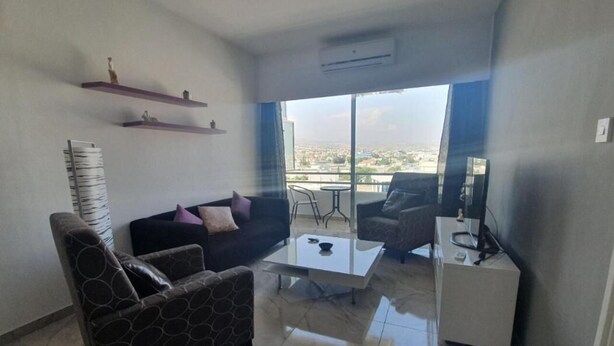 A modern living room with a sofa, armchairs, a coffee table, and a TV stand. There are two shelves with decorative items and a large window offering a city view. - Great views apartment. (Limassol)