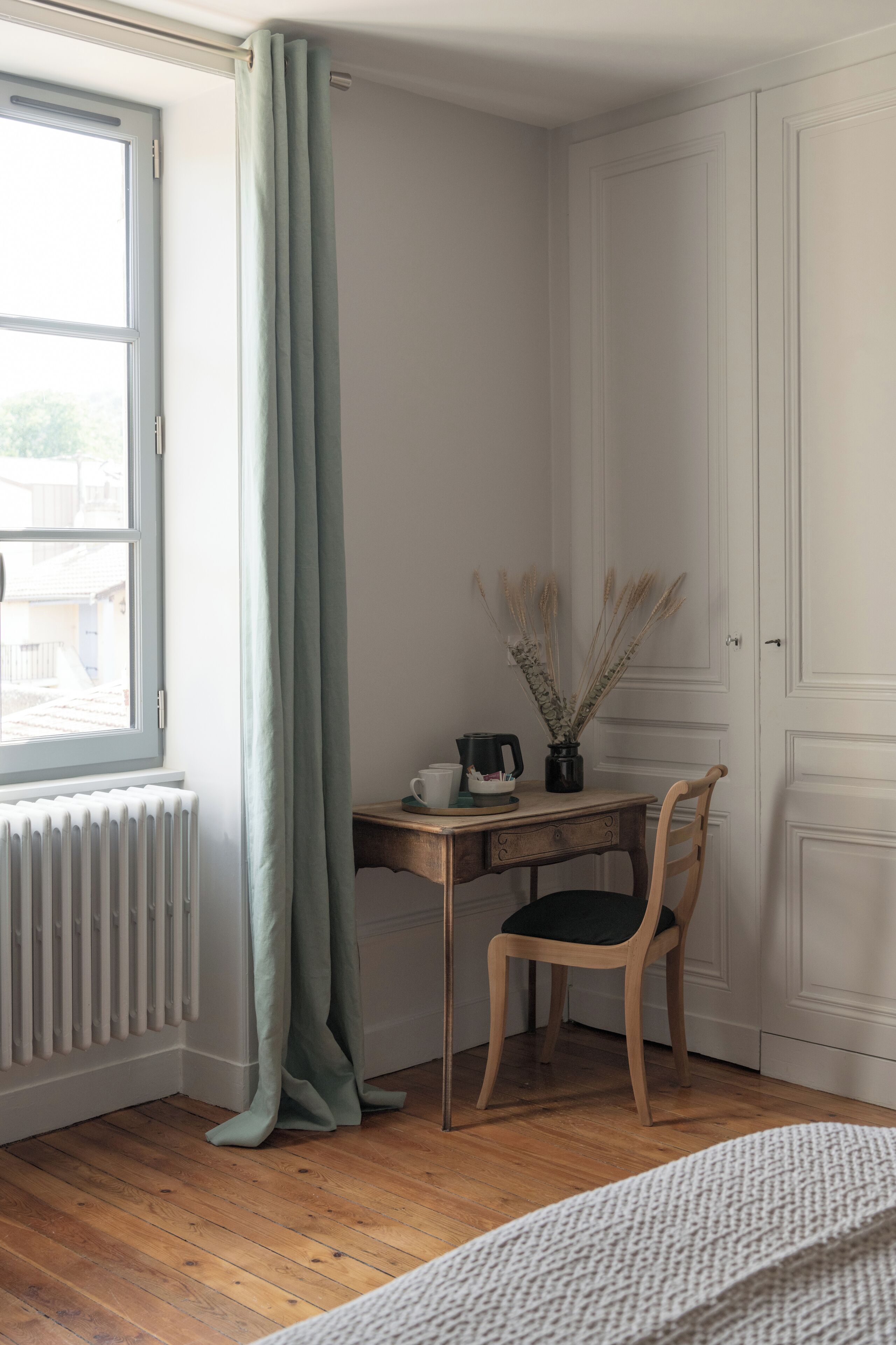 Ein Zimmer mit Schreibtisch aus Holz, einem Sessel, einem Heizkörper, einem Fenster mit Vorhängen und einer Tür.