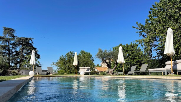A clear pool with lounge chairs and umbrellas, surrounded by trees and a house in the background. - La Maison des Monts d'or (Saint-Germain-au-Mont-d'Or)