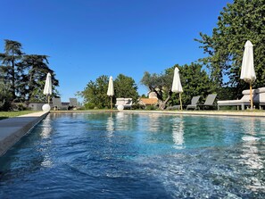 A clear pool with lounge chairs and umbrellas, surrounded by trees and a house in the background. - La Maison des Monts d'or (Saint-Germain-au-Mont-d'Or)