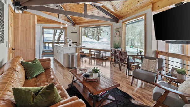 Amplia sala de estar con techo de madera, un ventanal grande con vistas a un paisaje nevado, un sofá cómodo con cojines, una mesa de centro de madera y un comedor con sillas y una mesa.