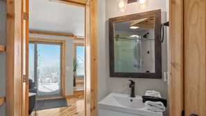 Un baño moderno con suelo de madera, un lavabo blanco, un espejo grande y vistas a un paisaje nevado a través de la puerta.