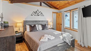 Un dormitorio con techo de madera, una cama con colcha gris, un televisor montado en la pared y una ventana con cortinas.