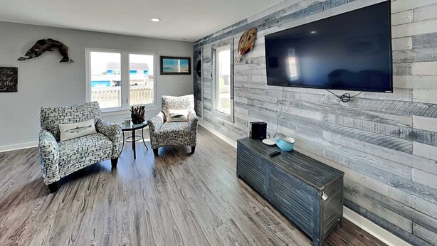 Una sala de estar con dos sillones estampados, una mesa de centro, un televisor de pantalla plana montado en una pared de madera y un ventanal con vistas al exterior.