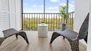 A balcony with wicker chairs, a small round table, and potted plants. - Admirals Bay 152 | Canal Front Condo, Balcony + Community Pool (Fort Myers Beach)