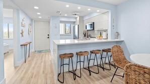 A modern kitchen with a white island, bar stools, and a woven chair. - Admirals Bay 152 | Canal Front Condo, Balcony + Community Pool (Fort Myers Beach)