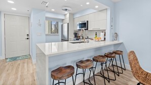 A modern kitchen with a white island, bar stools, and a woven chair. - Admirals Bay 152 | Canal Front Condo, Balcony + Community Pool (Fort Myers Beach)