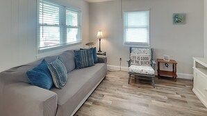 Un salon avec un canapé gris, des coussins bleus, une chaise en bois ornée d’un coussin à motifs de plage, une petite table en bois et une lampe.