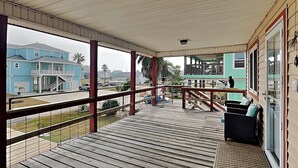 Une terrasse extérieure couverte, dotée d’un plancher en bois, d’une rambarde en métal et offrant une vue sur un quartier résidentiel avec des maisons et des palmiers.