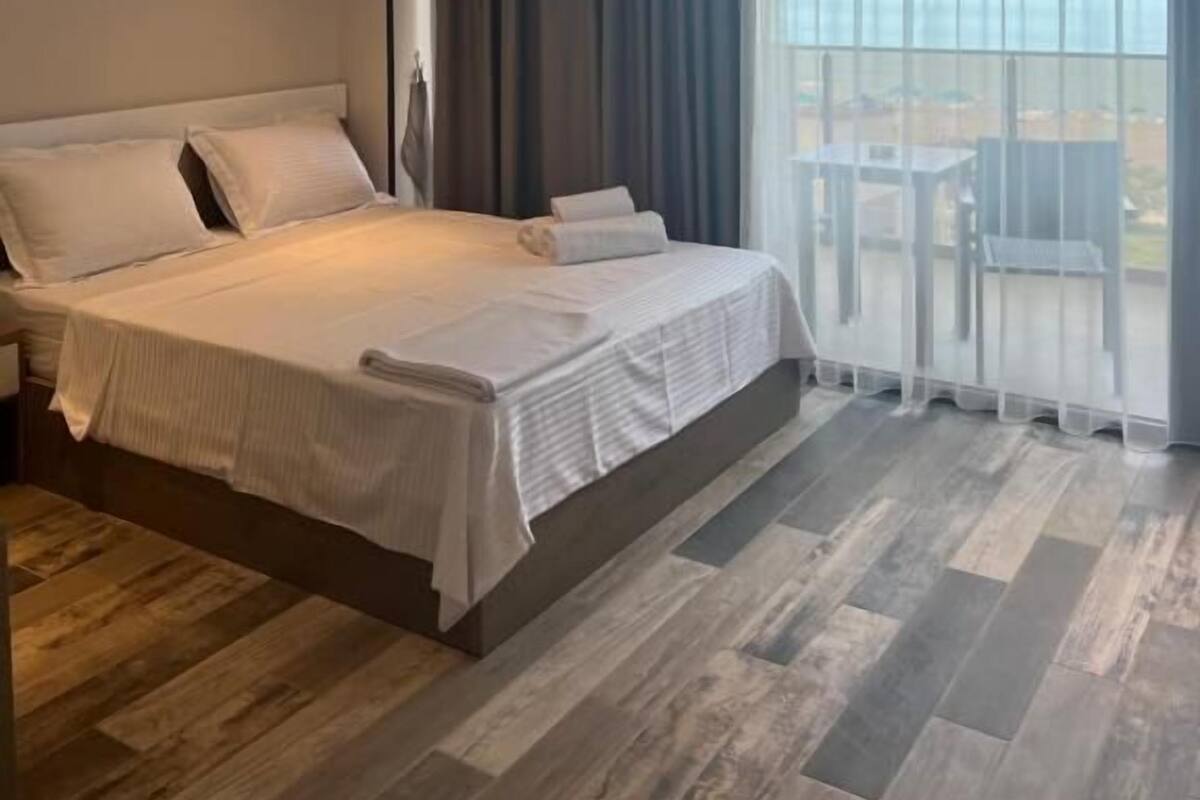 A hotel room with a large bed, a view of the sea, and a balcony with outdoor furniture.