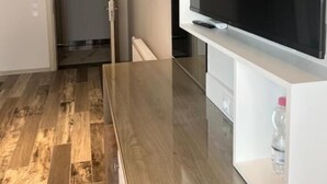 Ein modernes Wohnzimmer mit einem großen Flachbildfernseher, der an einem weißen Schrank befestigt ist, Holzboden und einer Tür zu einem anderen Raum.