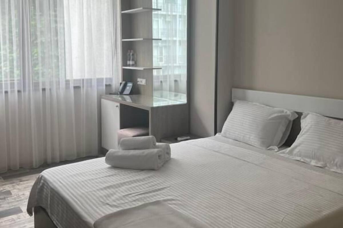 A modern hotel room with a large bed, a bedside table, a built-in shelf unit, and a window with sheer curtains.