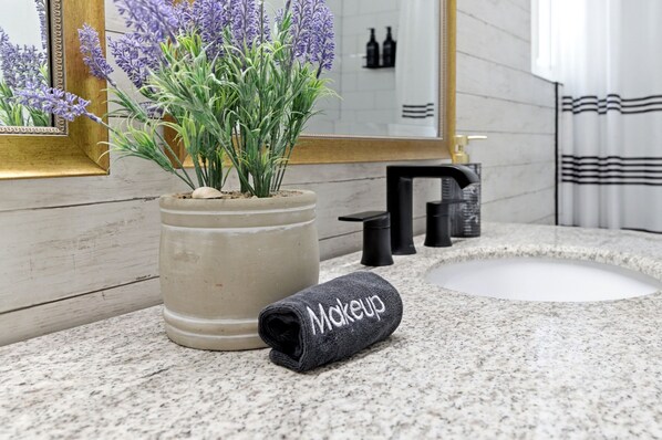Un baño con encimera de mármol, un lavamanos, una planta de lavanda en maceta, una toalla con el bordado 'Makeup' y un espejo con marco dorado.