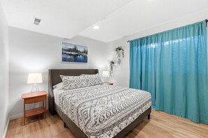 Une chambre à coucher avec un lit, une table de chevet, une plante fixée au mur, un tableau et une fenêtre avec des rideaux.
