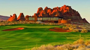 Un edificio moderno con fachada de vidrio, ubicado en un campo de golf bien cuidado con formaciones rocosas rojas al fondo.