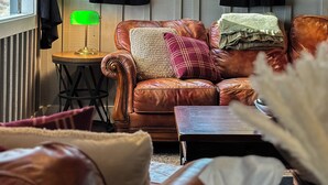 A cozy living room with a leather sofa, a small side table with a lamp, and a view of trees through a window. - The Duck Blind at Cooter Creek Cabins (Elberton)