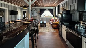 A kitchen with a central island, bar stools, a sofa, a television, and a window with curtains. - The Duck Blind at Cooter Creek Cabins (Elberton)