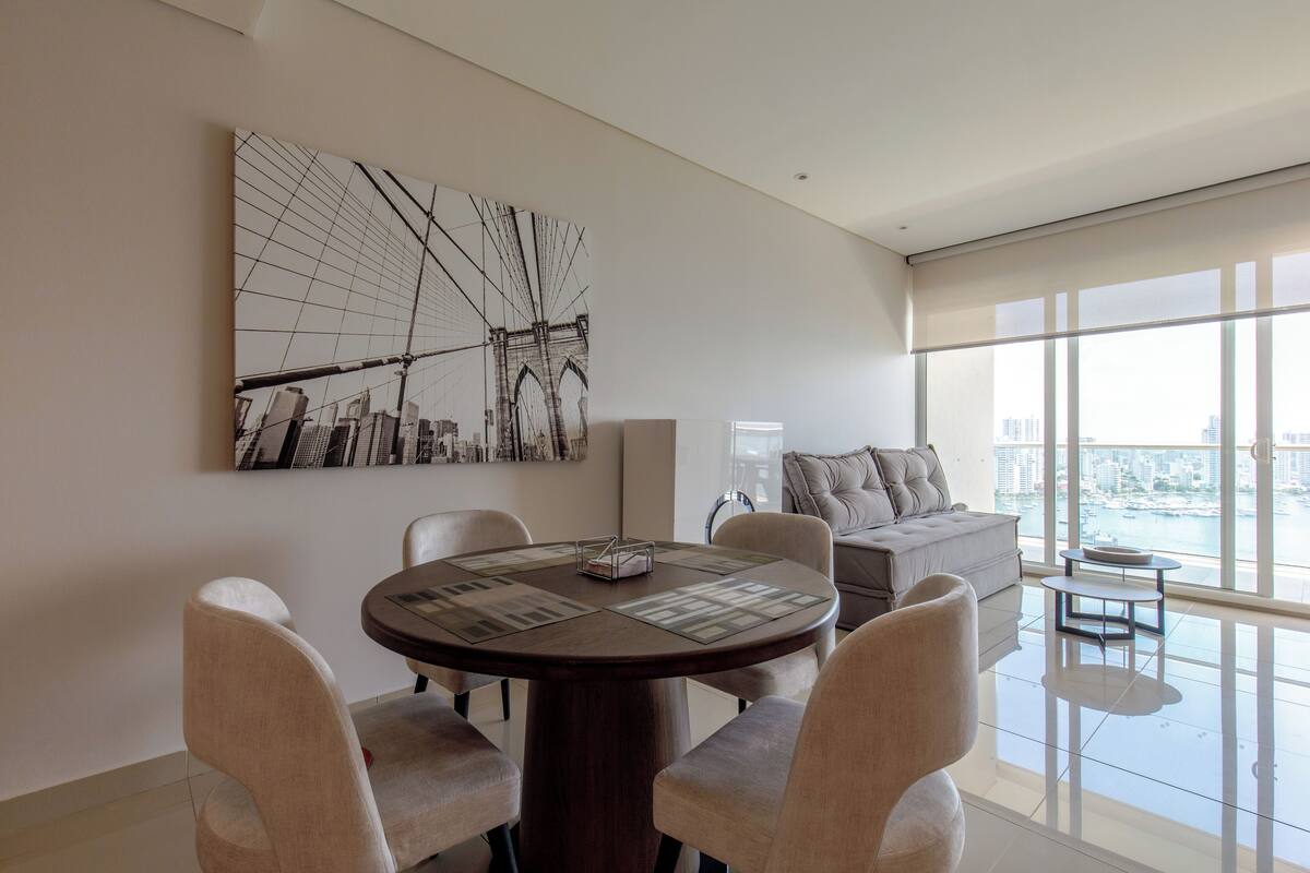 A modern living room with a round dining table, beige chairs, a large abstract painting, a sofa, and a view of the city through large windows.