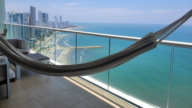 A balcony with a view of a beach and ocean, a hammock, and modern city buildings in the background. -  H2 Bocagrande Vista al Mar-Bahía (Cartagena)