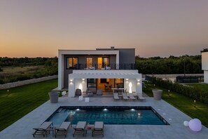 Una casa moderna de dos pisos con piscina, mobiliario exterior y un césped cuidado.