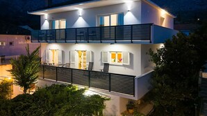A two-story modern house with a balcony, large windows, and a mountain backdrop. - Great 2-Bedroom Holiday Apartment with terrace and sea-view! (Orebic)