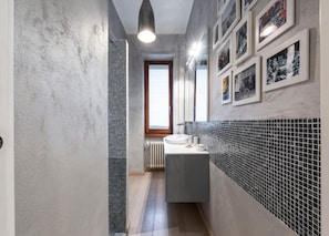 A narrow bathroom with a tiled backsplash, a sink, and a window with blinds.