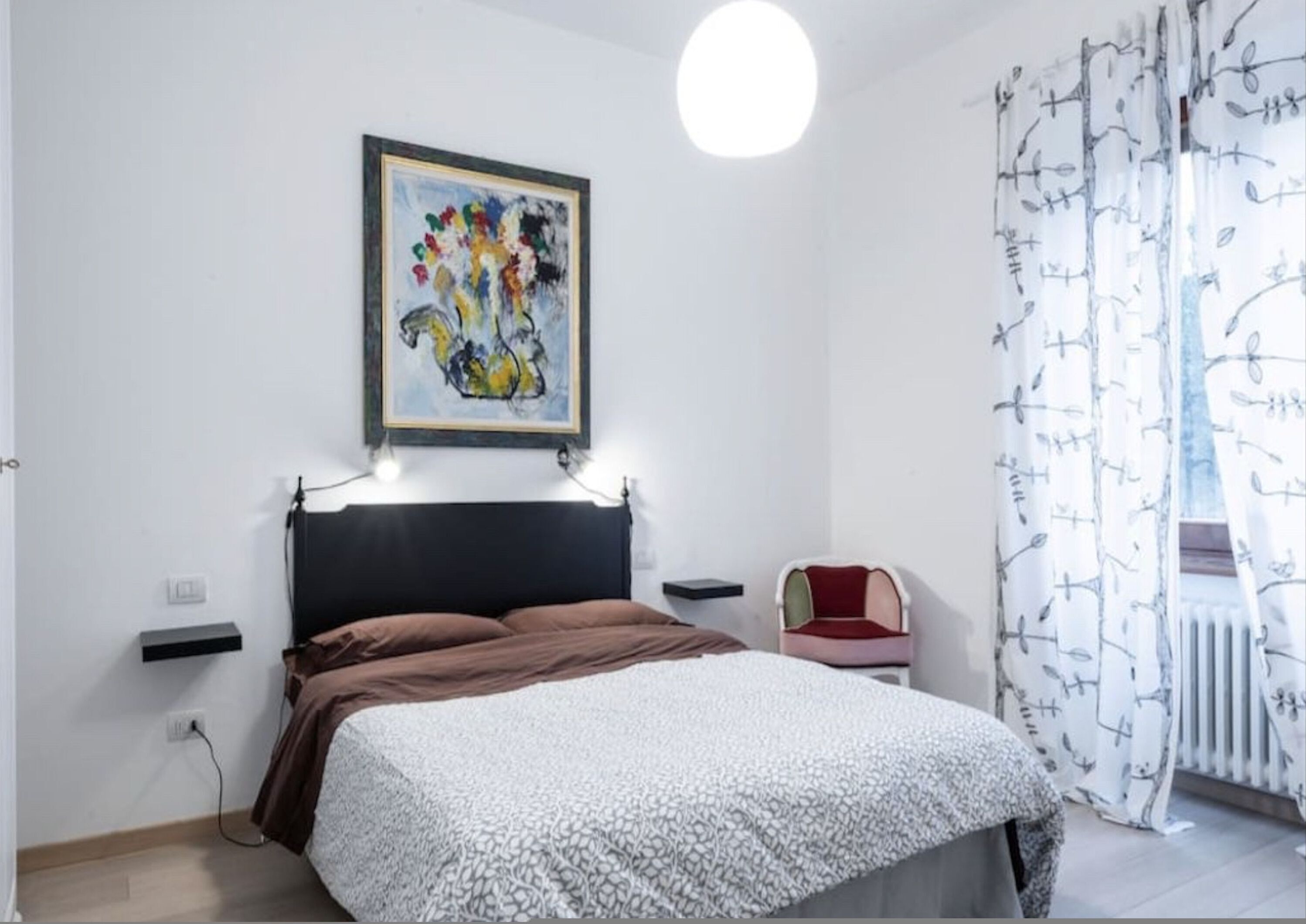 A bedroom with a bed, a nightstand, a chair, a radiator, and a painting on the wall.