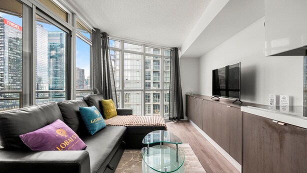 A modern living room with a sofa, glass coffee table, and a TV on a wooden cabinet. Large windows offer a city view. - GLOBALSTAY 1 Bedroom with a Romantic View of Trains in Downtown (Toronto/Canada)