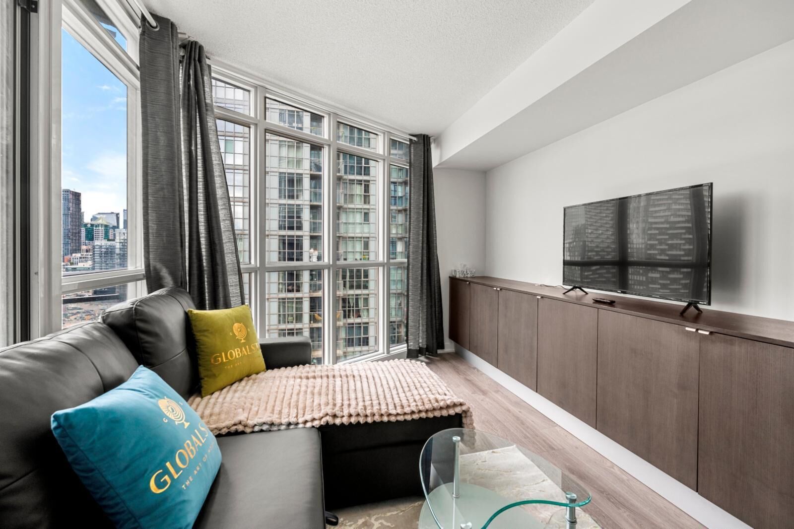 A modern living room with a large window, a sofa with cushions, a glass coffee table, and a TV mounted on the wall.