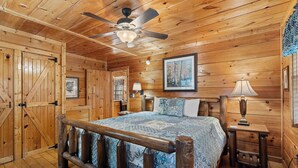 A wooden cabin bedroom with a bed, a ceiling fan, a nightstand, and a framed picture on the wall.