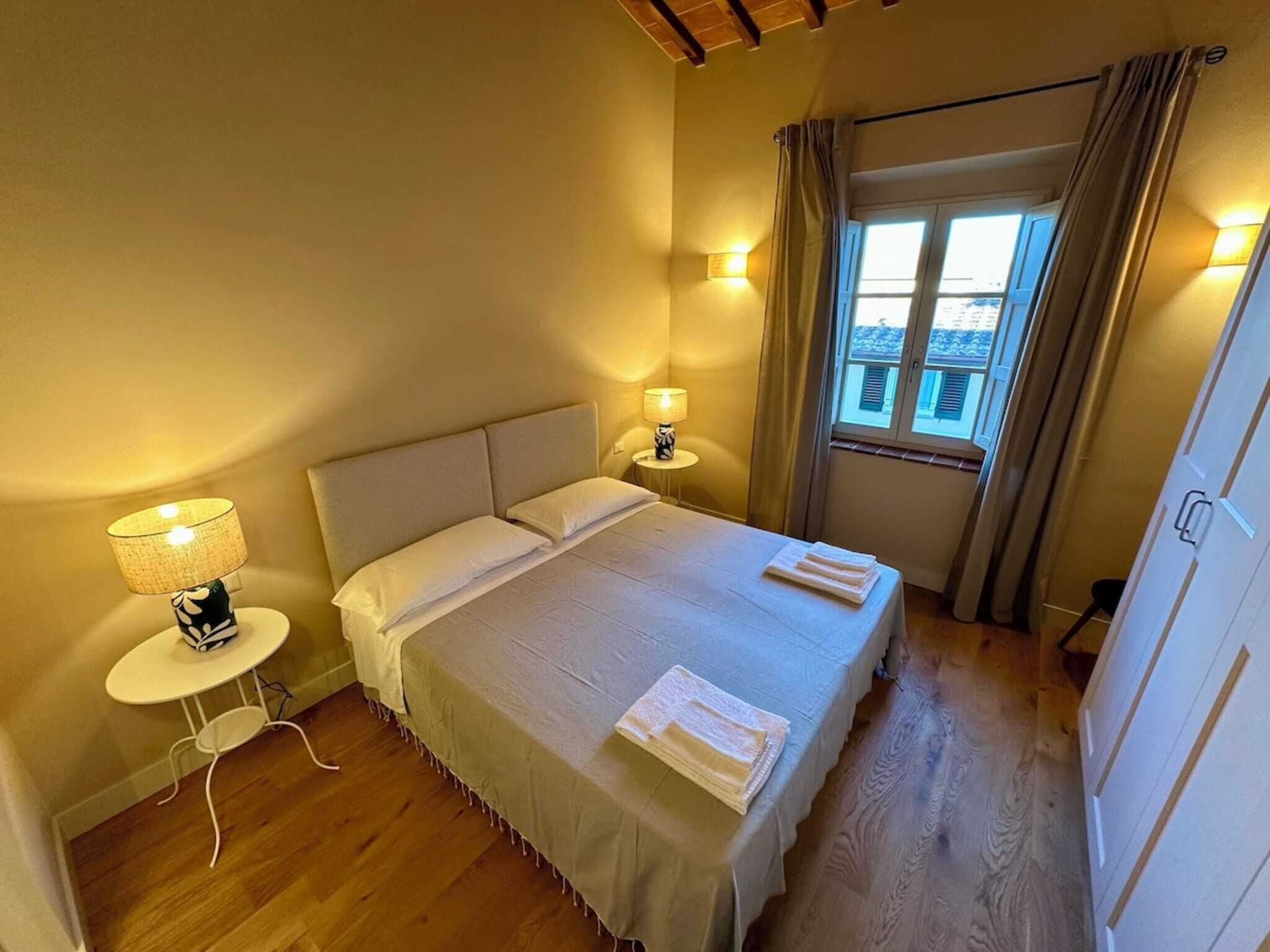 A neatly made bed with pillows and a blanket, a bedside table with a lamp, and a window with curtains.